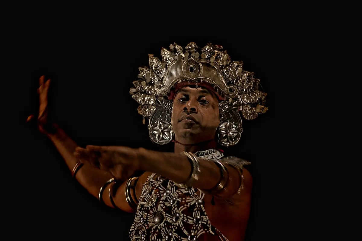 A male Kandyan dancer against a black background, wearing a metal-fronted headdress with silver ear plates and a beaded chest ornament, one arm extended