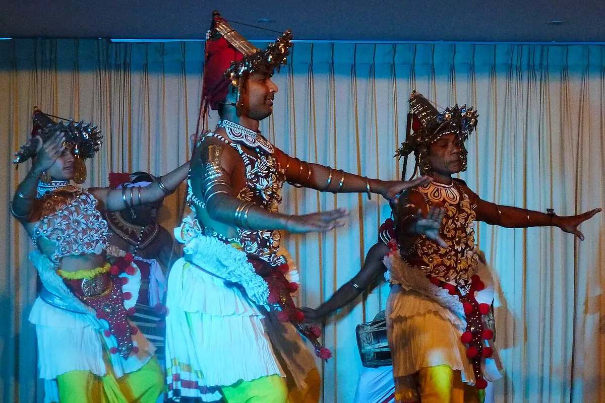 Three Kandyan dancers on a stage in beaded chest pieces, white pleated skirts and metal-fronted headdresses, arms outstretched, in front of a gold curtain